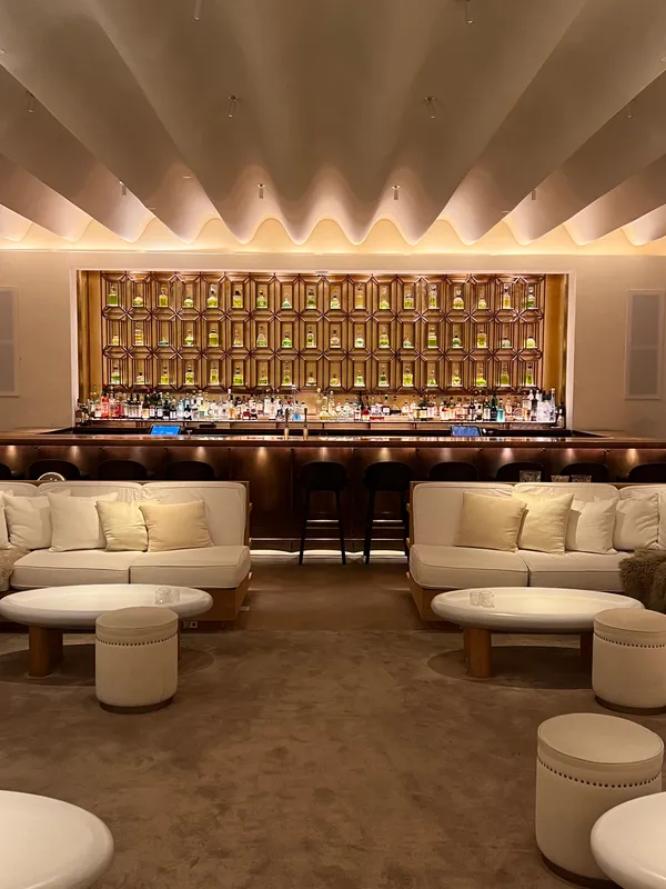 A hotel bar lounge with white sofas, a softly lit ceiling and a backlit wall of bottles.