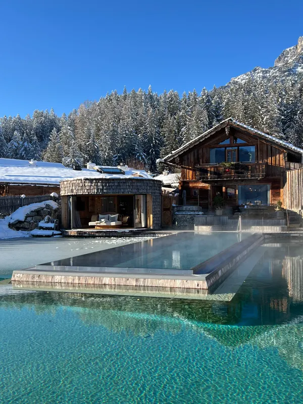 A heated outdoor pool in front of an alpine chalet, with snow-covered forest behind.